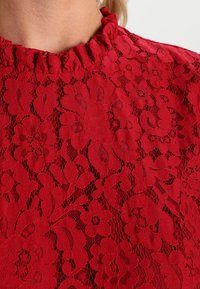 Punainen kukkakuvioinen pitsikangas, johon kuuluu teksturoitu muotoilu rypytetyllä kaula-aukolla, esittelee monimutkaisia kuvioita ja yksityiskohtaisia ompeluja.