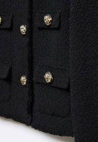 Veste en tissu noir texturé avec quatre boutons ronds dorés et deux poches à rabat, montrant un gros plan détaillé de la partie avant.