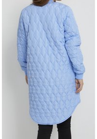 Henkilö, jolla on päällään pitkä, vaaleansininen tikattu takki, jossa on timantinmuotoiset tikit ja resorit hihansuissa, esitettynä selin yksinkertaista taustaa vasten.