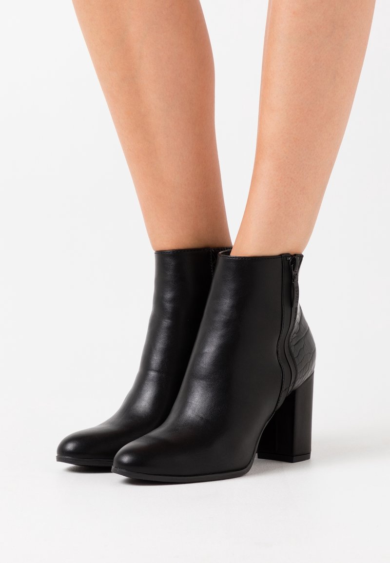 Bottines en cuir noir avec talons blocs et fermetures éclair latérales portées sur des jambes nues sur fond blanc.