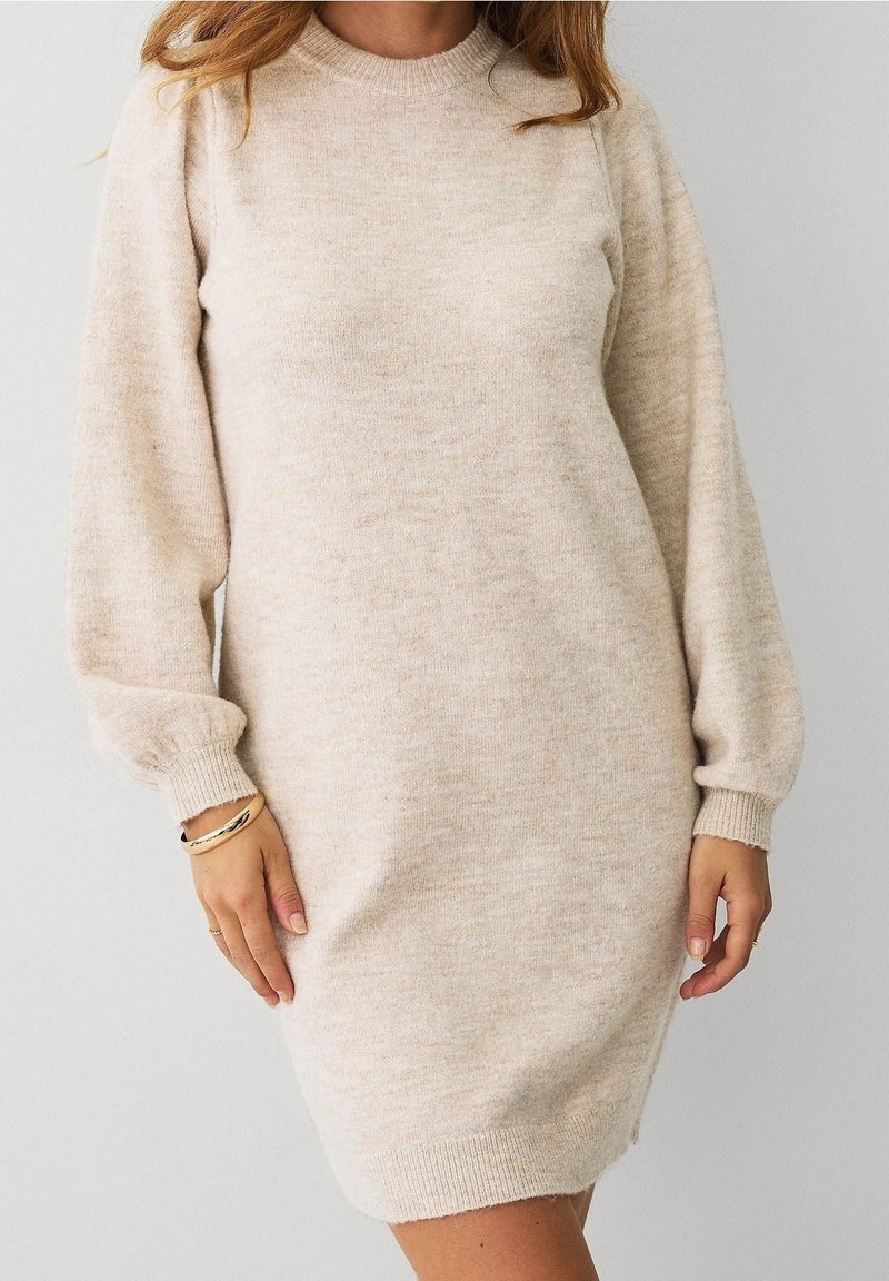 Femme portant une robe-pull en tricot beige à manches longues avec poignets et ourlet côtelés, debout devant un fond clair uni.