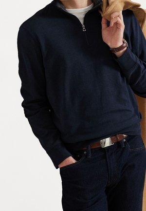 Homme portant un pull marine à fermeture éclair quart, un maillot blanc, un jean foncé, une ceinture en cuir marron et une montre, tenant un manteau camel sur l'épaule.