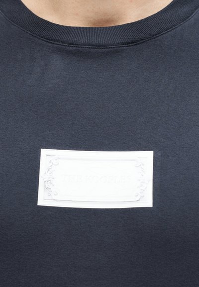 Κοντινό πλάνο σκούρου γκρι μπλουζάκι με στρογγυλή λαιμόκοψη και λευκή ανάγλυφη ορθογώνια ετικέτα στο στήθος με το λογότυπο "THE KOOPLES".