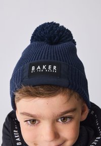 Tengerészkék bordázott kötött sapka bojttal a tetején és fekete címkével, amelyen a "BAKER BY TED BAKER" felirat olvasható. Puha tapintású, testhez simuló kialakítás.