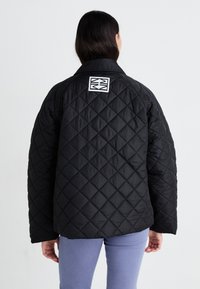 Veste noire matelassée avec un motif en losanges, manches larges et grand col. Un logo carré est visible sur le haut du dos.