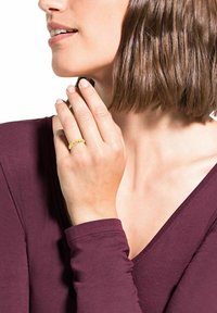 Kvinna i en vinröd topp som rör vid sitt haka medan hon håller ett litet föremål, bärande en gul flätad ring på sin finger. Naturligt ljus framhäver hennes drag.