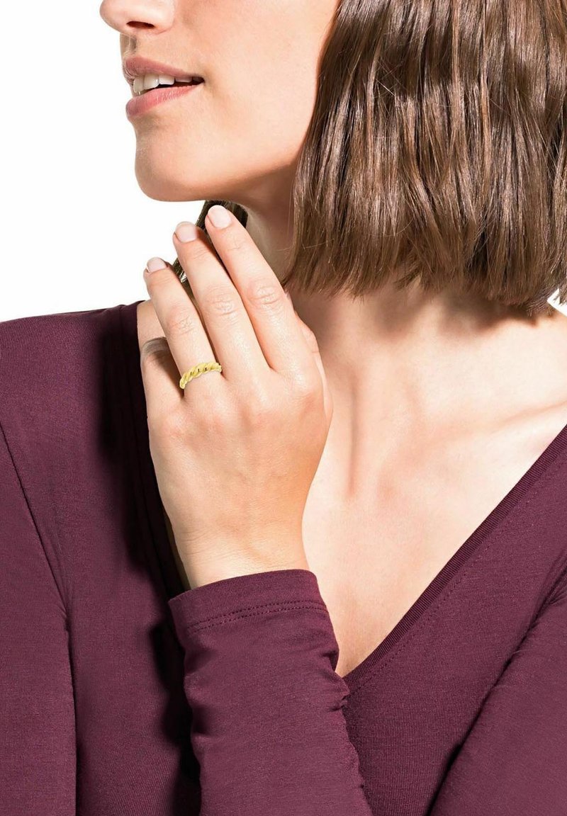 Kvinna i en vinröd topp som rör vid sitt haka medan hon håller ett litet föremål, bärande en gul flätad ring på sin finger. Naturligt ljus framhäver hennes drag.