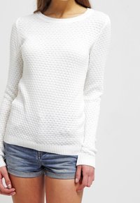 Vit stickad tröja med en texturerad bikakmönster och ribbade ärmslut, kombinerad med jeansshorts. Tröjan har en rund halsdesign.