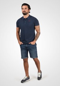 Mørkeblå polo-shirt med korte ærmer, denimshorts og grå sneakers. Har et logo på skjorten og optrukne manchetter på shortsene.