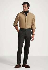 Άνδρας φορώντας μπεζ πουκάμισο με κουμπιά πάνω από μαύρο λαιμόκοψη, σκούρες γκρι προσαρμοσμένα παντελόνια, καφέ λευκά παπούτσια με φουντίτσες και ρολόι χειρός, στέκεται με το ένα χέρι στην τσέπη.