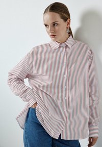 Camicia a maniche lunghe con bottoni di colore bianco a righe verticali rosse. Presenta un colletto a punta e una vestibilità comoda; abbinata a jeans blu.