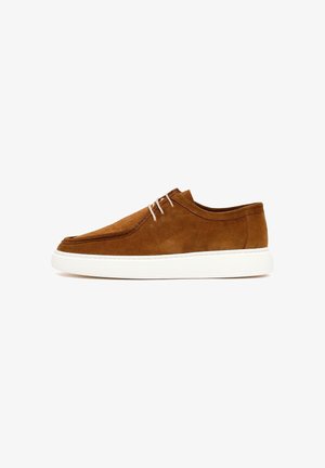 Bruine suède casual schoen met witte rubberen zool en lichtbeige veters, zijaanzicht op een witte achtergrond.