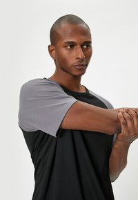 Sort og grå sportstrøje med korte ærmer. De grå skuldre har et struktureret design, mens kroppen er glat og tætsiddende.