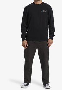 Sudadera negra con cuello redondo y mangas largas, con un pequeño logo en el pecho. Combina con pantalones oscuros y zapatillas negras y blancas.