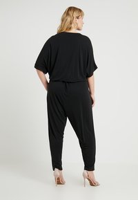 Svart jumpsuit i lett stoff, med løs passform, vide ermer og smale ben. Et bemerkelsesverdig trekk er den stramme midjen.
