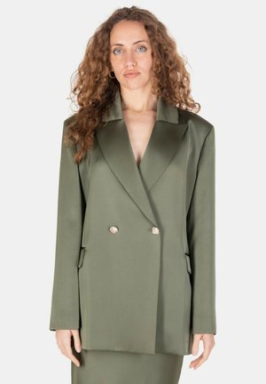 Donna con capelli ricci che indossa un blazer verde oliva oversize con bottoni dorati su sfondo semplice.