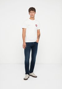 Camiseta blanca de algodón con gráfico bordado en el lado izquierdo del pecho, combinada con jeans ajustados azul oscuro y zapatillas blancas con suelas marrones.