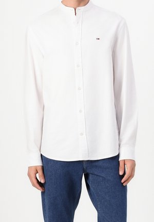 Mężczyzna w białej, zapinanej na guziki, koszuli z długim rękawem i kołnierzykiem mandaryńskim oraz w niebieskich dżinsach, stojący na białym tle.