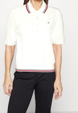 Vrouw draagt een wit poloshirt met korte mouwen, met een kraag en zoom met rode, witte en zwarte strepen, gecombineerd met een zwarte broek.