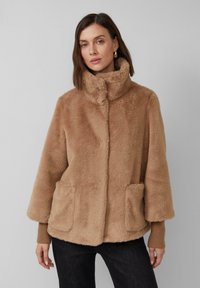 Manteau en fausse fourrure couleur brun clair, avec un col montant, deux poches avant et des poignets en tricot côtelé. Texture douce et coupe décontractée.