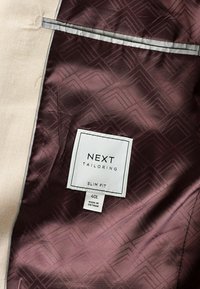 Forro de chaqueta ajustada en satén burdeos con patrón geométrico. La etiqueta dice "NEXT TAILORING," talla 40L, fabricada en Vietnam.