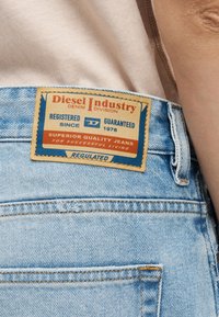 Κοντινό πλάνο της πίσω τσέπης ανοιχτόχρωμου τζιν με μια δερμάτινη ετικέτα Diesel Industry Denim Division σε κόκκινες, μπλε και ταμπά αποχρώσεις.