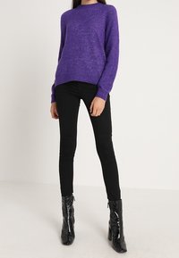 Lila gestrickter Pullover mit rundem Ausschnitt und langen Ärmeln, kombiniert mit schwarzen engen Hosen und schwarzen, glänzenden Stiefeletten.