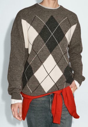 Personne portant un pull marron à motif losange argyle blanc et noir, une chemise blanche en dessous, et un pull rouge noué autour de la taille.