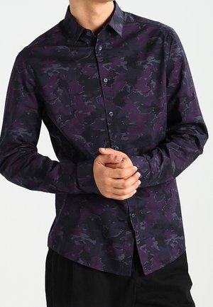 Homme portant une chemise à manches longues boutonnée avec un motif violet foncé et noir, les mains jointes devant.