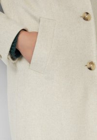 Manteau en laine beige clair avec une seule rangée de grands boutons, une poche latérale et une texture douce. Présente un poignet replié avec un intérieur vert.