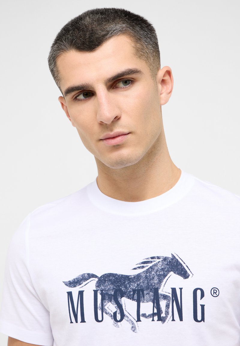 T-shirt blanc à manches courtes avec un graphique bleu d'un cheval au galop et le mot "MUSTANG" en texte navy en gras. Matière en coton doux.