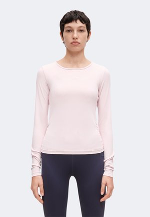 Persona con el pelo corto y oscuro lleva una camiseta de manga larga rosa claro y mallas oscuras, de pie de frente contra un fondo claro y liso.