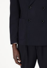 Blazer doppiopetto blu navy in tessuto testurizzato, con tre bottoni e una tasca. Le maniche terminano al polso, rivelando il polsino di una camicia bianca.