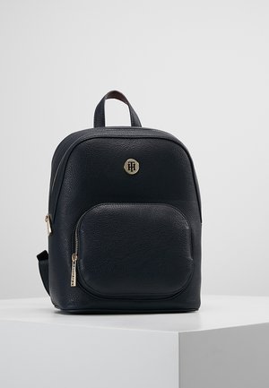 Zwarte rugzak van getextureerd leer met voorvak met ritssluiting, gouden ritstrekkers, bovenhandvat en ronde gouden logo-embleem op een gladde achtergrond.