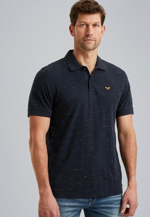 Polo shirt navy con colletto corto, texture sottile e un piccolo emblema dorato sul petto; abbinato a jeans in denim.