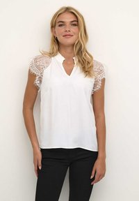 Blouse blanche avec un col et des manches courtes en dentelle. Elle présente une texture lisse et une coupe légèrement décontractée, complétée par des détails en dentelle.