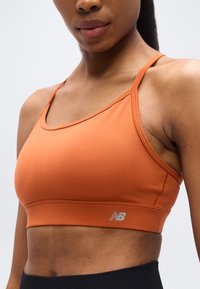 Πορτοκαλί αθλητικό σουτιέν με εφαρμοστή σχεδίαση, κατασκευασμένο από λείο ύφασμα. Διαθέτει λεπτές τιράντες και μικρό γκρι λογότυπο New Balance στη ζώνη.