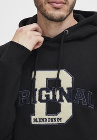 Black hoodie featuring textured cream and navy embroidery with the word "ORIGINAL" and "BLIND DENIM" on the front. Drawstring hood visible.