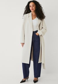 Lang cremet frakke med klapkrave over en hvid top, parret med mørkeblå høj talje jeans og sorte spidse sko.