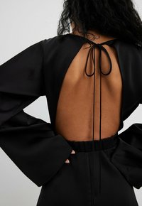 Robe noire avec de longues manches cloche et un dos ouvert, présentant une fermeture à nouer. Tissu lisse avec une taille froncée.