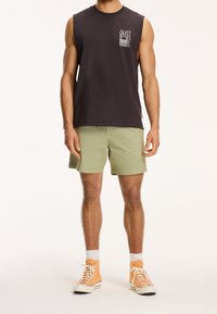 Tmavé tílko bez rukávů s designem grafické kapsy na přední straně, spárované se světle zelenými šortkami a oranžovými vysokými teniskami, které mají bílé tkaničky.