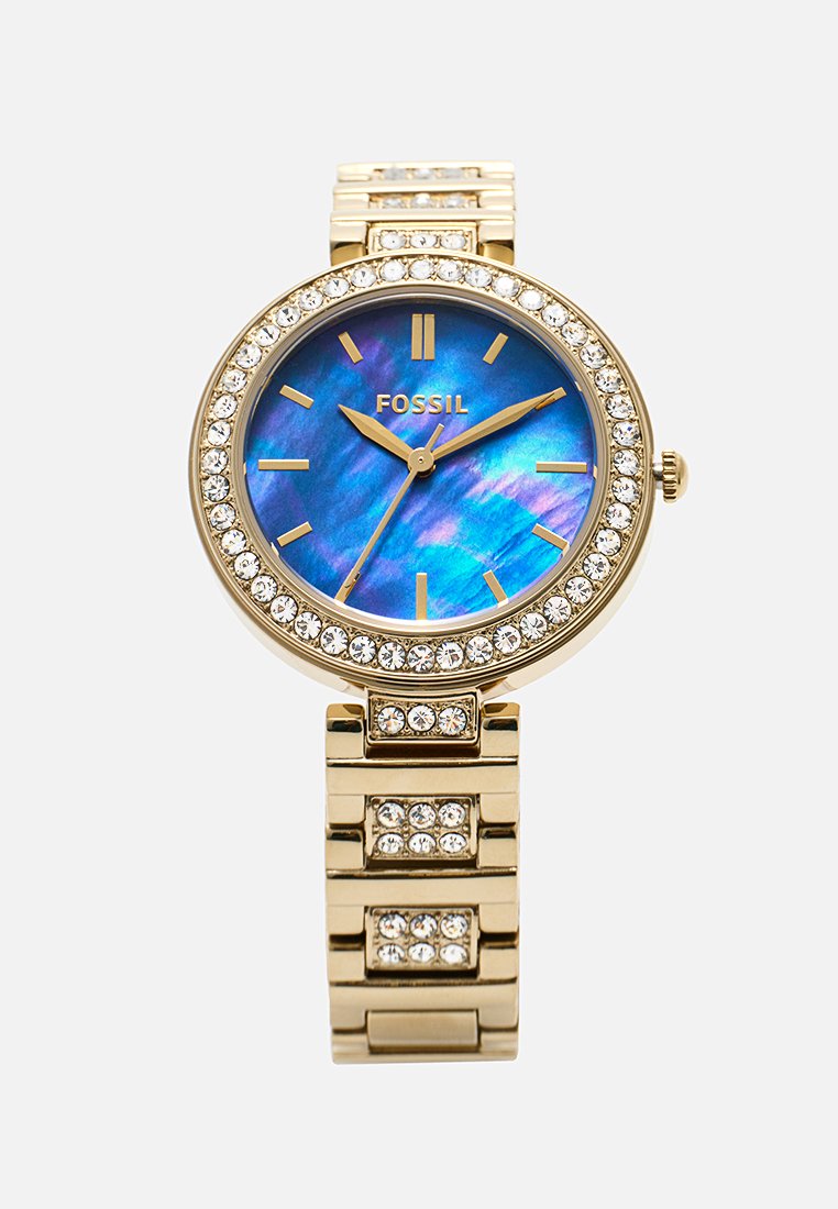 Montre dorée avec un cadran rond, un cadran en bleu irisé, une lunette en cristal et un bracelet en métal à maillons ornés d'accents décoratifs.