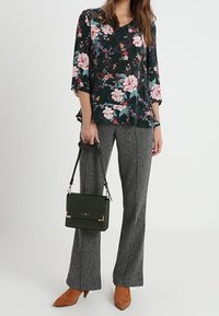 Blus med blommönster i mörkgrön färg, med trekvartslånga ärmar och volangkant, matchad med grå texturerade byxor och en grön handväska.