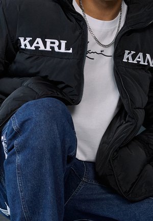 Personne portant un jean bleu, un t-shirt blanc avec un motif de ligne noire, une doudoune noire avec le texte "KARL" et un collier chaîne argenté.