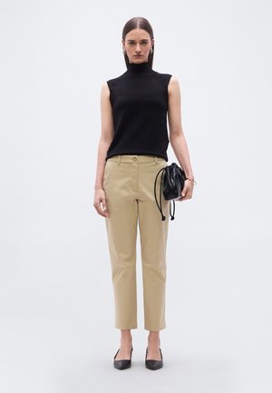 Moteris, dėvinti juodą berankovį golfą, smėlio spalvos kelnes, juodus aukštakulnius, rankose laikanti juodą krepšį su raišteliu, stovinti priešais paprastą pilką foną.