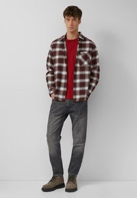 Młody mężczyzna stoi, ubrany w czerwoną koszulkę, rozpiętą brązową i białą koszulę w kratę, wyblakłe czarne dżinsy i brązowe buty na sznurowadła, na tle szarej ściany.