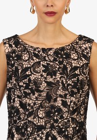 Abito in pizzo nero con motivi floreali su una fodera color nudo, caratterizzato da un collo rotondo e design senza maniche. Orecchini a cerchio dorati completano il look.