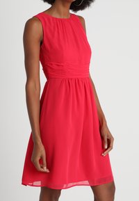 Robe rouge sans manches avec une taille froncée, un tissu fluide et une texture douce et légère. Longueur genou avec un col rond.