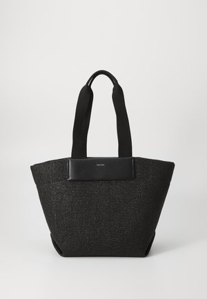Sac de toile noir tissé avec de larges poignées en tissu et un petit patch rectangulaire en cuir arborant un logo blanc "Calvin Klein".