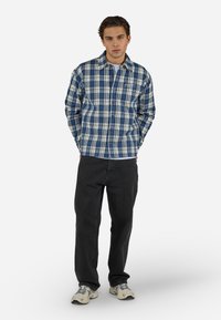 Jeune homme debout, les mains dans les poches, portant une chemise à carreaux bleue et blanche, un pantalon noir et des baskets beiges sur fond blanc.